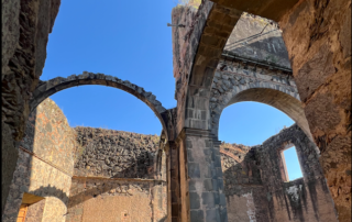 Nombrado Pueblo Mágico desde 2015, Mascota, Jalisco, cautiva a quienes lo visitan con su aire puro, sus paisajes encantadores y su fascinante historia. Entre sus numerosos atractivos destaca la Iglesia Inconclusa de La Preciosa Sangre de Cristo. Ubicado en el corazón del pueblo, en las calles Libertad y Rosa Dávalos, este imponente lugar, con sus arcos majestuosos y estructuras incompletas, transporta a los visitantes al siglo XIX. El templo fue concebido en 1896 para ser el Santuario de la Virgen de Talpa y las obras, realizadas completamente a mano, avanzaron hasta 1904 cuando los movimientos pre-revolucionarios que estallaron en 1905 dificultaron su culminación. En 1908, el gobierno incautó los bienes destinados a su construcción para financiar la guerra, interrumpiendo los trabajos y posteriormente, en 1926, el lugar fue tomado por los cristeros durante el conflicto religioso, y las paredes incompletas se convirtieron en escenarios de encarcelamientos y fusilamientos. Tras esos oscuros episodios, la construcción quedó abandonada y en 1945, el templo fue declarado patrimonio nacional, lo que aseguró su preservación como una reliquia histórica. Hoy, sus arcos y torres inacabadas permanecen como un testimonio del pasado, inmortalizadas en el tiempo. Actualmente, estas ruinas son uno de los puntos más visitados de Mascota, su imponente arquitectura y el peso de su historia cautivan a quienes se acercan a conocerlas. Si decides visitar este mágico lugar, no olvides dejar una contribución económica al entrar, lo recaudado se destina al mantenimiento del sitio, para que las generaciones futuras puedan seguir admirándolo.